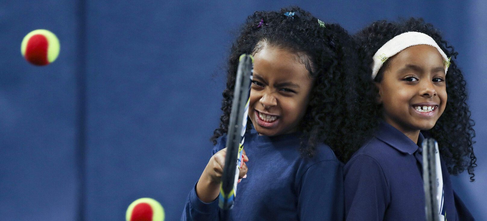 Two dark - skinned young female - presenting child holding tennis racquets and smiling.
