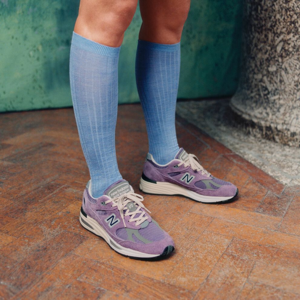 A pair of New Balance Made in UK sneakers standing on a wooden floor with textured green walls and a stone pillar in the background.