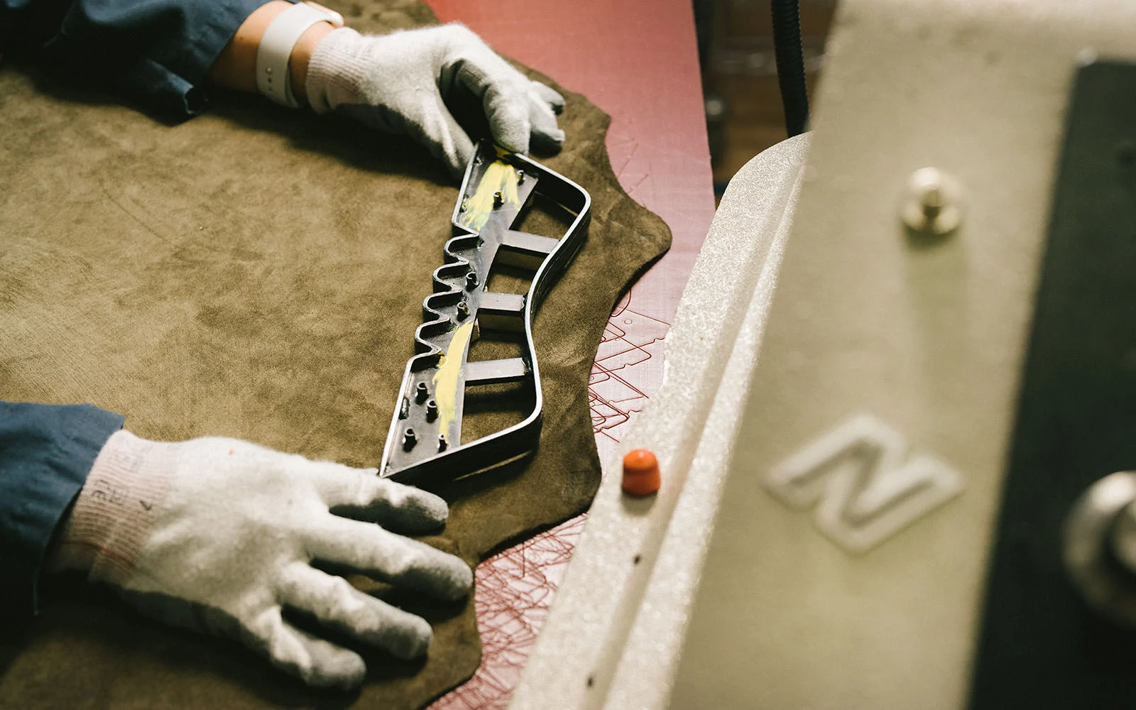 A pair of hands laying a tool over a piece of suede in a factory setting. 