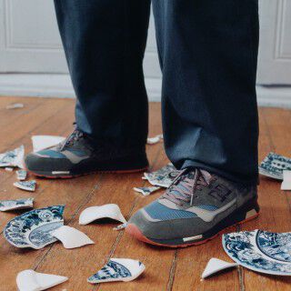 Knee down view of a man wearing a pair of New Balance Made in UK sneakers standing on a rustic wooden floor with smashed blue and white ceramic crockery all over the ground and white cabinets in the background. 