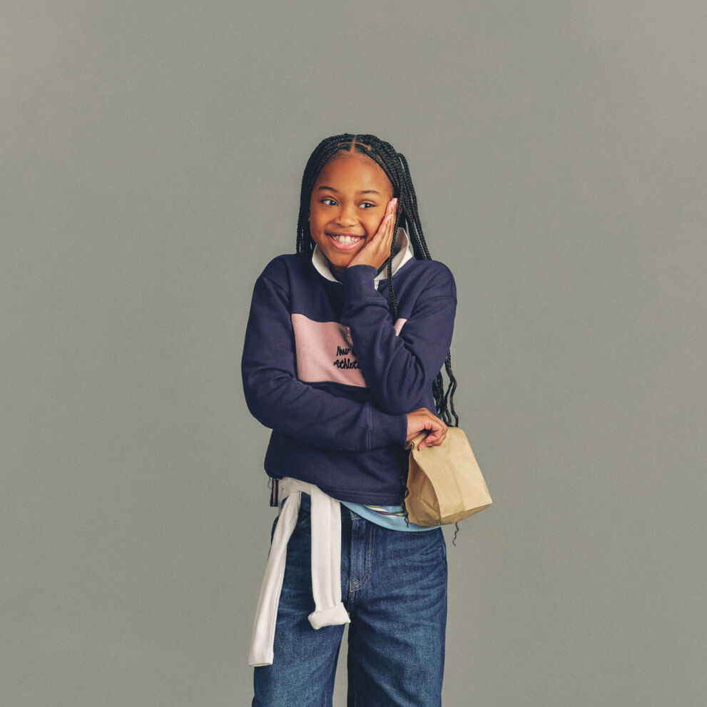 The child is wearing a light gray T-shirt layered under a soft pink zip-up hoodie with a white New Balance logo on the chest.